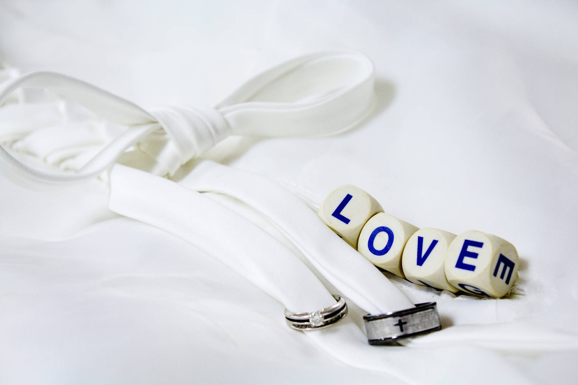 Cuvântul „Love” scris pe patru zaruri albe, lângă două verighete argintii și negre legate cu o panglică albă, așezate pe o foaie albă. — Dragoste Fundal
