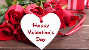 Buchet de trandafiri roșii așezat pe masă, cu un cartonas în formă de inimă albă pe care scrie "Happy Valentine's Day". — Chromebook Fundal