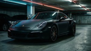 Un Porsche 911 Turbo S negru mat iese în evidență într-un garaj subteran sumbru, cu reflexii de neon. imagine de fundal